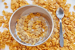 Cornflakes in a metal bowl with milk on a painted white wooden background. Cornflakes scattered on a wooden table with spoon.