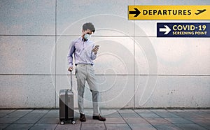 Corona Virus Situation Concept. Young Passenger Businessman wearing Surgical Mask. Using Smartphone. Standing with Luggage in the