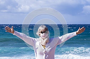 Coronavirus. Attractive senior woman with wind in her hair enjoying beauty of the sea, wearing a protective mask against