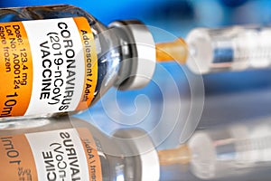 Coronavirus covid-19 vaccine concept -  glass bottle with silver cap on glossy white desk, orange syringe injected,  blurred blue
