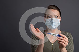 Coronavirus, a girl in a mask holds a temperature meter with a high temperature, on a gray background. Headlines for media, news.