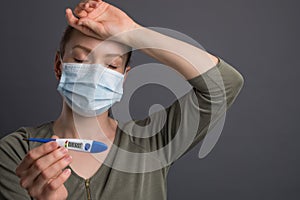 Coronavirus, a girl in a mask holds a temperature meter with a high temperature, on a gray background. Headlines for media, news.