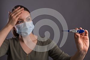 Coronavirus, a girl in a mask holds a temperature meter with a high temperature, on a gray background. Headlines for media, news.