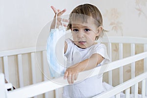 Coronavirus lockdown covid-19  concept, precautions. little cute toddler stands in the crib at home and holds out a medical mask