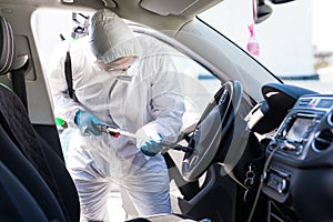 Coronavirus Pandemic. Disinfector in a protective suit and mask sprays disinfectants of car outdoors