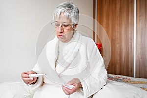 Coronavirus. Sick senior woman holding tissue and thermometer sit on bed, upset old mature woman caught cold got