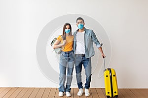 Coronavirus and tourism. Young couple in medical masks ready for vacation during pandemic, standing with baggage