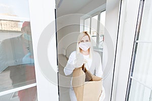 Coronavirus. Woman wearing a medical mask and rubber gloves receiving a package from a delivery man indoors. Virus