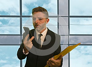 Corporate company work lifestyle portrait of stressed and frustrated businessman looking at mobile phone holding paperwork reports