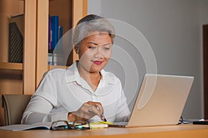 Corporate job lifestyle portrait of happy and successful attractive middle aged Asian woman working at office laptop computer desk