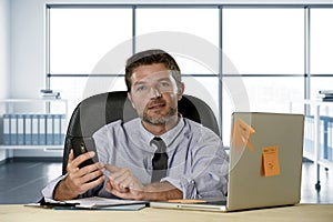 Corporate portrait of happy successful businessman in shirt and tie smiling at computer desk with mobile phone
