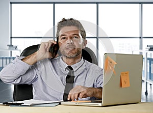 Corporate portrait of happy successful businessman in shirt and tie smiling at computer desk with mobile phone