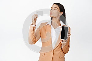 Corporate woman kissing her credit card, showing order on mobile phone screen, empty display on smartphone, standing in