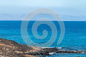 Corralejo along black rocks, blue water, view on volcanic Lobos island, Fuerteventura, Canary islands