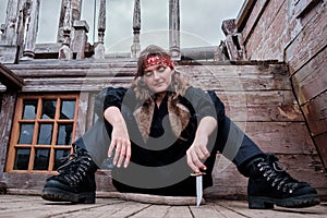 Corsair girl in a black clothes sits on the wooden floor of an retro pirate ship and draws a knife on the deck