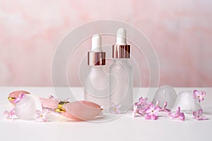 Cosmetic bottles of essential oil, face massage roller and lilac flowers on pink background. Aromatherapy, facial skin care