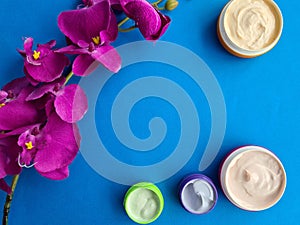 Cosmetic creams in jars with flowers on blue background copy space