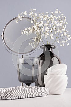 Cosmetic and make up accessories and home decoration black glass vase with flowers, silver mirror, bowls on soft white wood table.