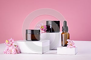 Cosmetic mockup -  jars for cream, essential oil bottle of amber glass on white podiums with pink spring flowers. Template.