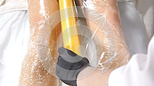 Cosmetic procedure body wraps. Beautician in black gloves covers with anti-cellulite cellophane female legs in modern