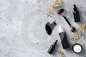 Cosmetic set of black obsidian face massage roller, gua sha, serum, moisturizer cream and golden branches on concrete background.