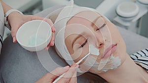 Cosmetician hands applying cream mask on young woman`s face at beauty spa salon