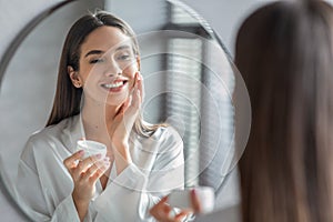 Cosmetics Concept. Young Woman Looking At Mirror And Applying Face Cream