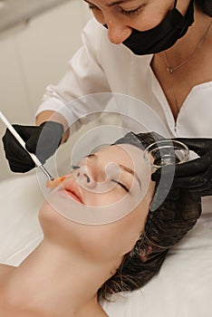 The cosmetologist applies a liquid gel to the face with a wide brush, the carboxytherapy procedure and its preparations.