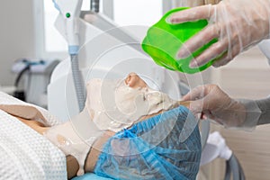 The cosmetologist applying an alginate mask to the face of a young lying woman in beauty salon for procedure of