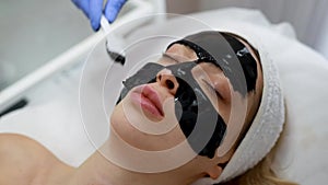 Cosmetologist applying black mask onto woman`s face in spa salon. Facial rejuvenation and cleansing procedure.