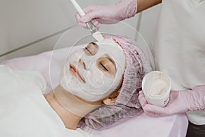 Cosmetologist applying mask on client& x27;s face in spa salon. Young woman getting facial care by beautician at spa salon Acne