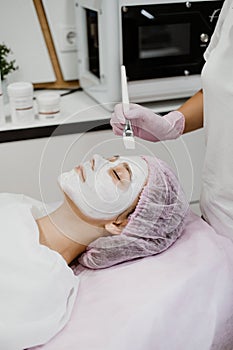 Cosmetologist applying mask on client& x27;s face in spa salon. Young woman getting facial care by beautician at spa salon Acne