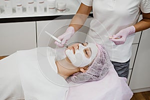 Cosmetologist applying mask on client& x27;s face in spa salon. Young woman getting facial care by beautician at spa salon Acne