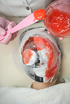 cosmetologist applying red alginate mask to face for her female client in beauty salon