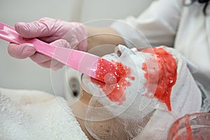 cosmetologist applying red alginate mask to face for her female client in beauty salon