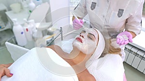 Cosmetologist is applying white mask on woman client face in beauty clinic.