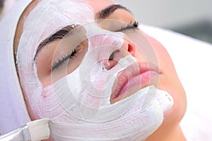 Cosmetologist is applying white mask on woman client face in beauty clinic.