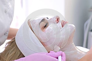 Cosmetologist is applying white mask on woman client face in beauty clinic.
