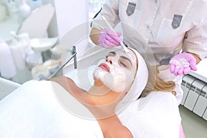 Cosmetologist is applying white mask on woman client face in beauty clinic.