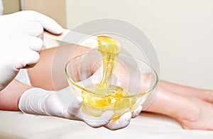 Cosmetologist beautician master holding bowl with hot wax for depilation epilation hair removal procedure and female legs on photo