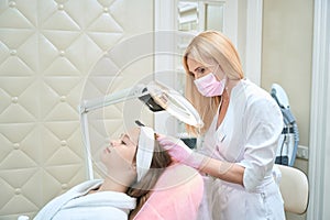 Cosmetologist checks girl head, looking through magnifying glass lamp