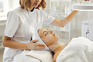 Cosmetologist or dermatologist directing lamp to womans face for facial treatment