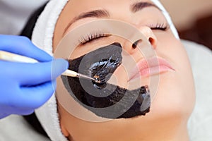 Cosmetologist does a black mask to cleanse the skin and anti-wrinkle on the face of a beautiful woman, spa treatments and photo