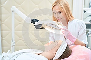 Cosmetologist looks through magnifying glass lamp at face of girl