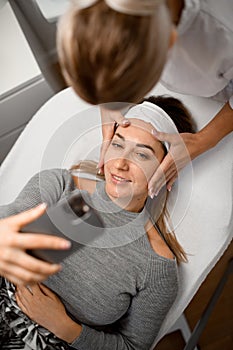 Cosmetologist makes a facial massage for a girl with fair skin who hold a mobile