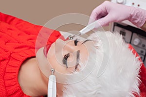 The cosmetologist makes the procedure of Microcurrent therapy of a beautiful, young woman in Santa Claus hat