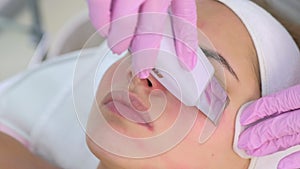 Cosmetologist makes ultrasonic face cleaning procedure to young woman in clinic.