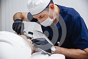 A cosmetologist male doctor in black gloves and blue uniform examines the patient& x27;s mole using a dermatoscope.