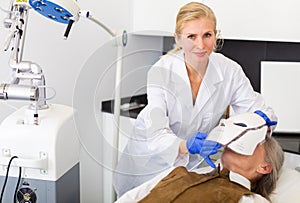 Cosmetologist performing anti-aging face phototherapy procedure for aged woman