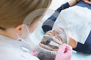 The cosmetologist in pink gloves with a brush applies a carbon mask for peeling on the face of a young girl in a